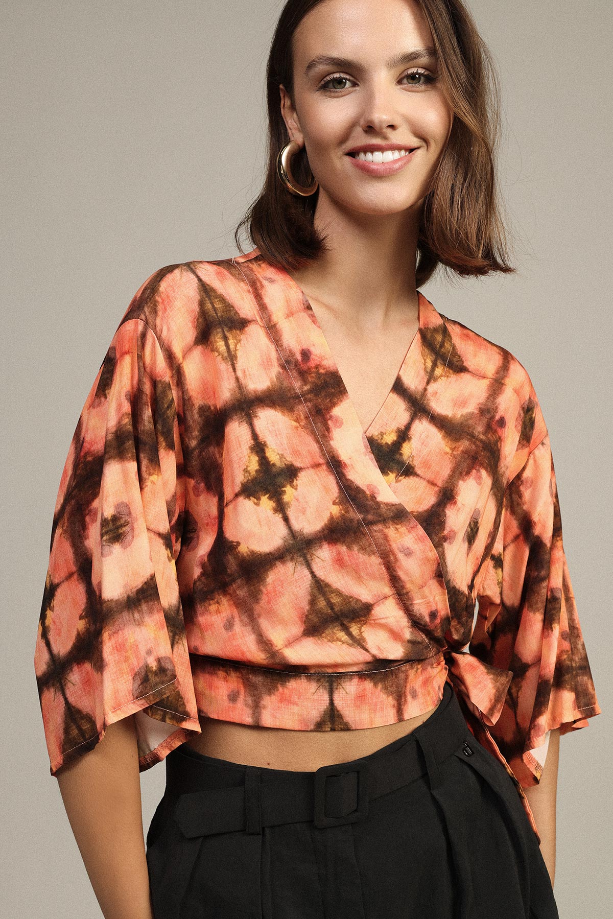 Smiling woman wearing orange and black tie-dye wrap crop top with wide sleeves and black high-waisted trousers
