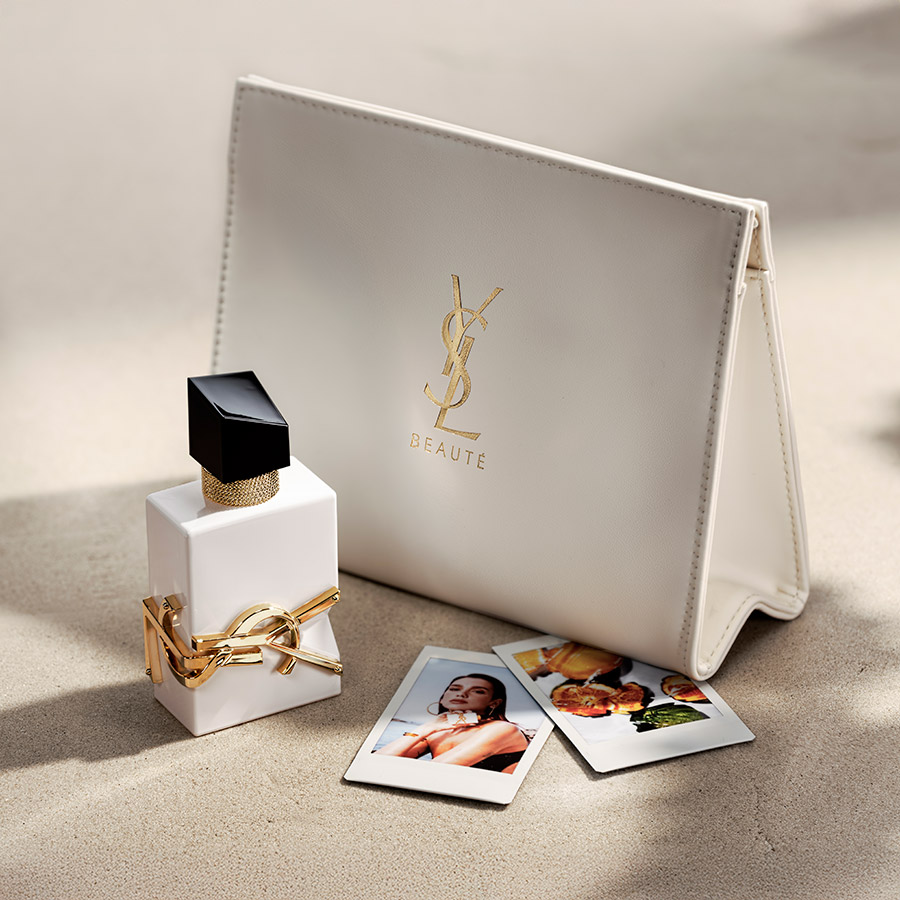 Un flacone di profumo bianco YSL Libre esposto accanto a una pochette in pelle bianca coordinata e foto polaroid sulla sabbia.