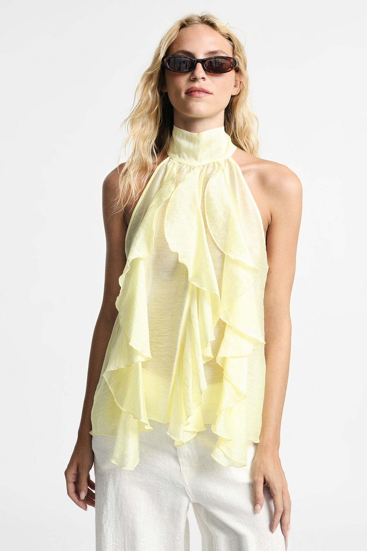 Woman wearing pale yellow ruffled halterneck top with white trousers and sunglasses, summer outfit