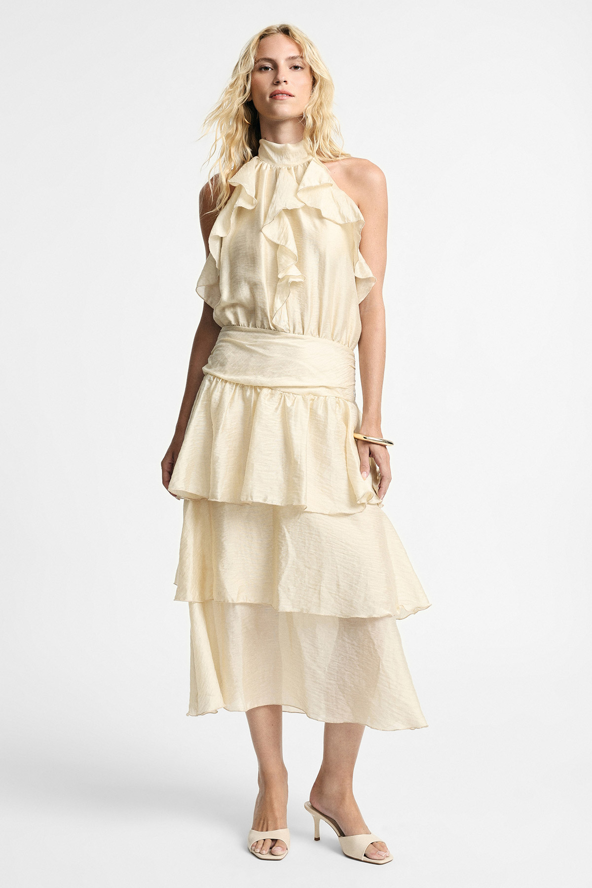 Woman wearing cream tiered ruffle halter midi dress with wide waistband and nude heeled mules on white background