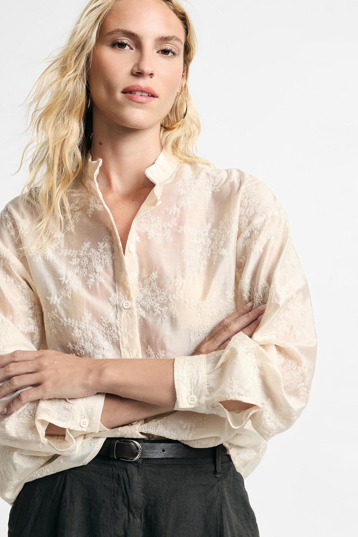 Blonde woman modelling a sheer cream embroidered blouse with rolled sleeves, black belt and dark trousers