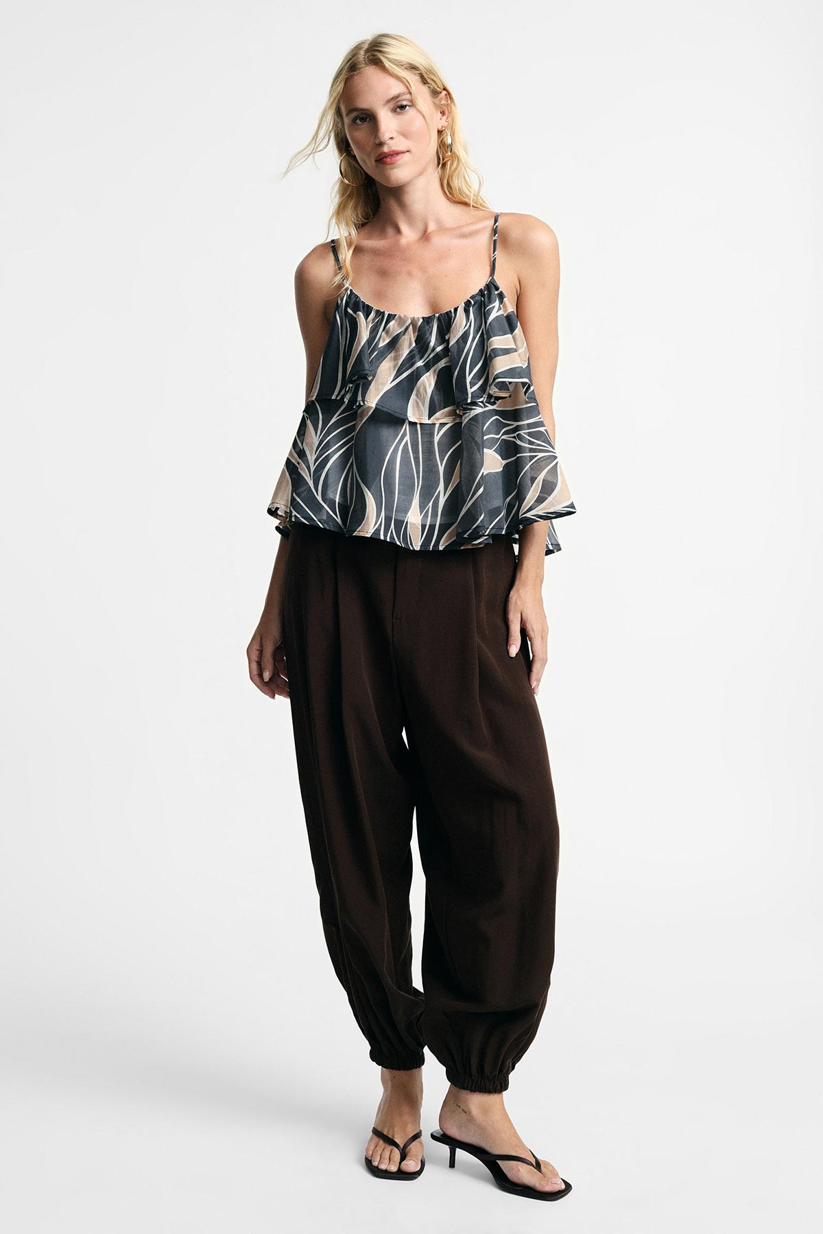 Model wearing patterned ruffle cami top with thin straps, loose dark harem trousers and black heeled sandals on white background