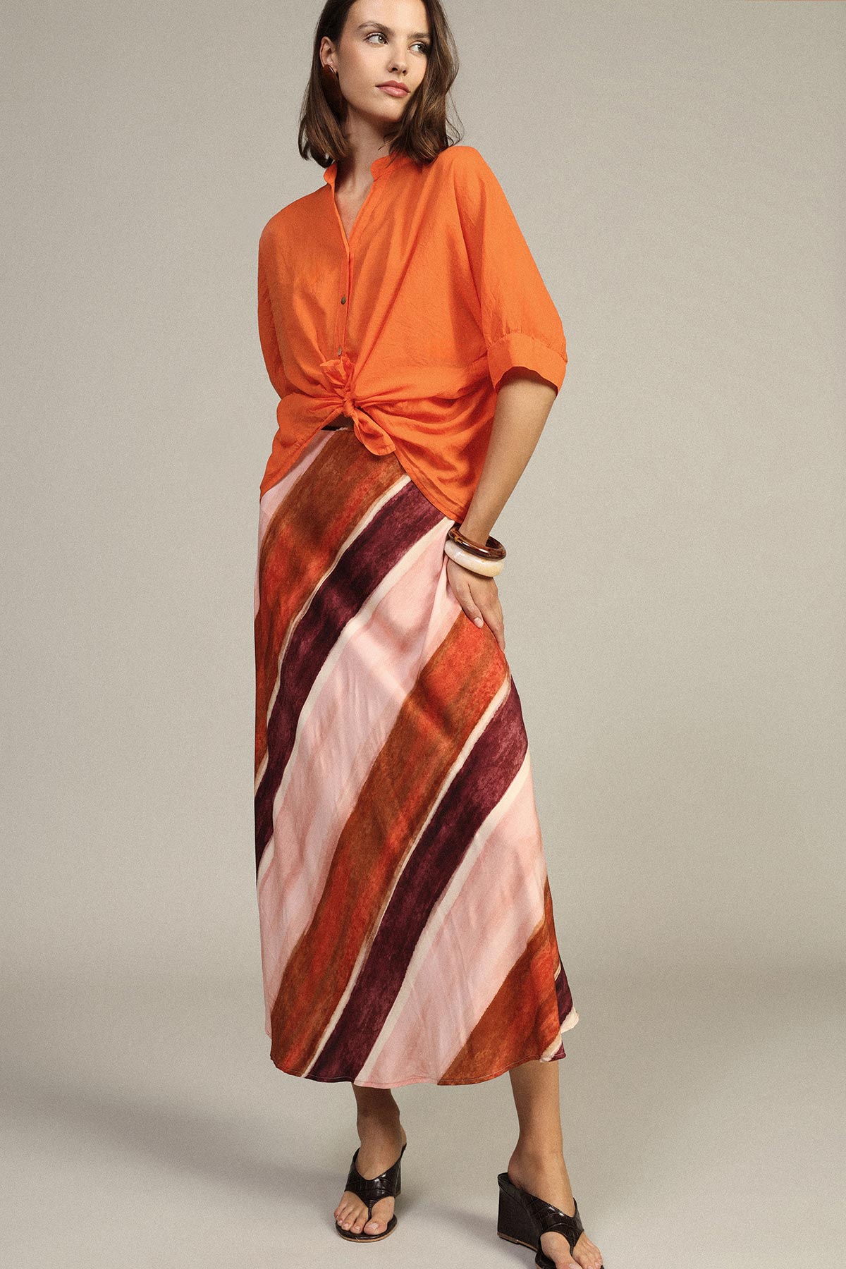 Woman wearing an orange tied blouse and diagonal striped pink, burgundy and rust maxi skirt with black sandals and bangles