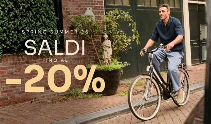 Man riding a bicycle past a brick storefront and potted statue; overlay reads 'Spring Summer 26 SALDI -20%' promoting men's summer fashion sale