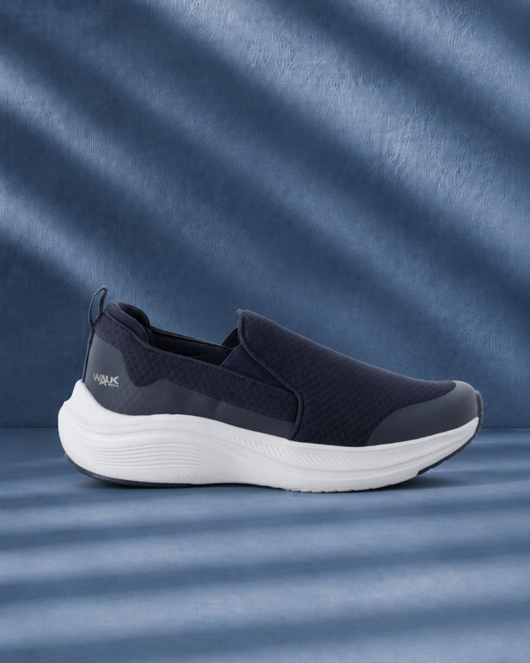 Navy slip-on athletic walking shoe with breathable mesh upper and white cushioned sole on textured blue background with shadow lines