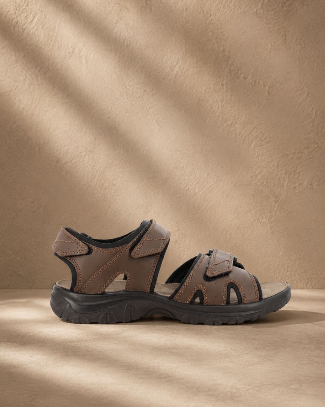 Brown leather men's outdoor sandal with adjustable Velcro straps on textured beige background