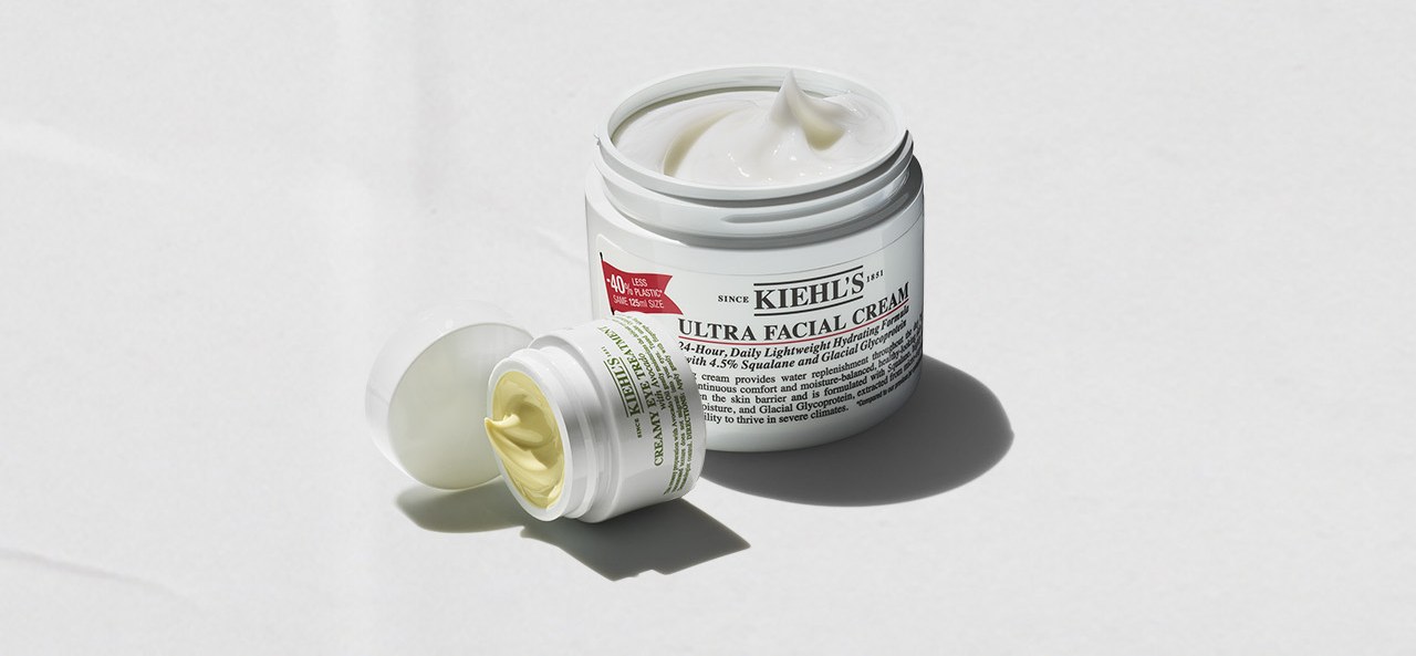 Vasetti aperti di Kiehl's Ultra Facial Cream e Creamy Eye Treatment con avocado su sfondo bianco
