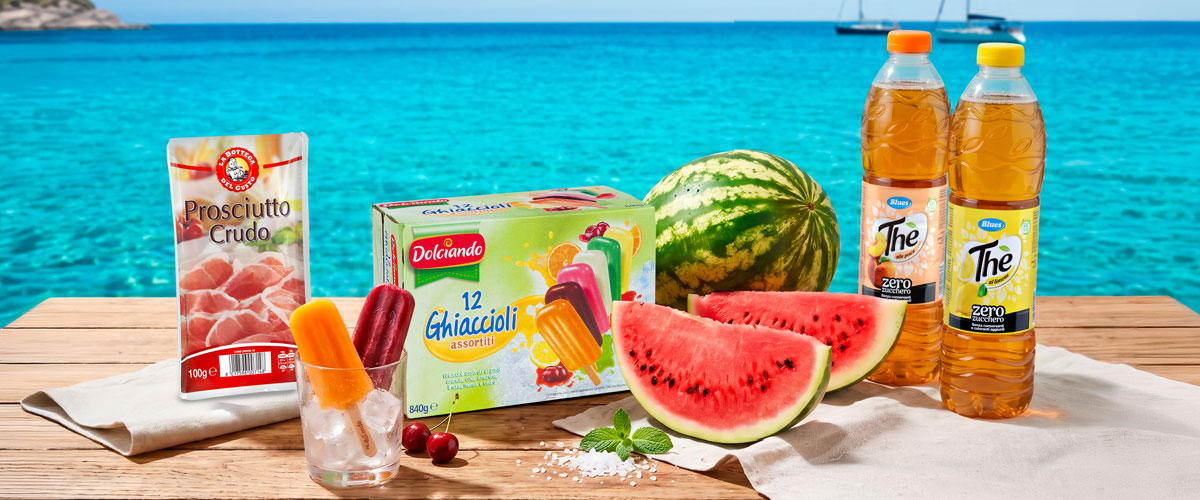 Seaside picnic with sliced watermelon, box of ice lollies, glass with popsicles on ice, prosciutto packet and iced tea bottles