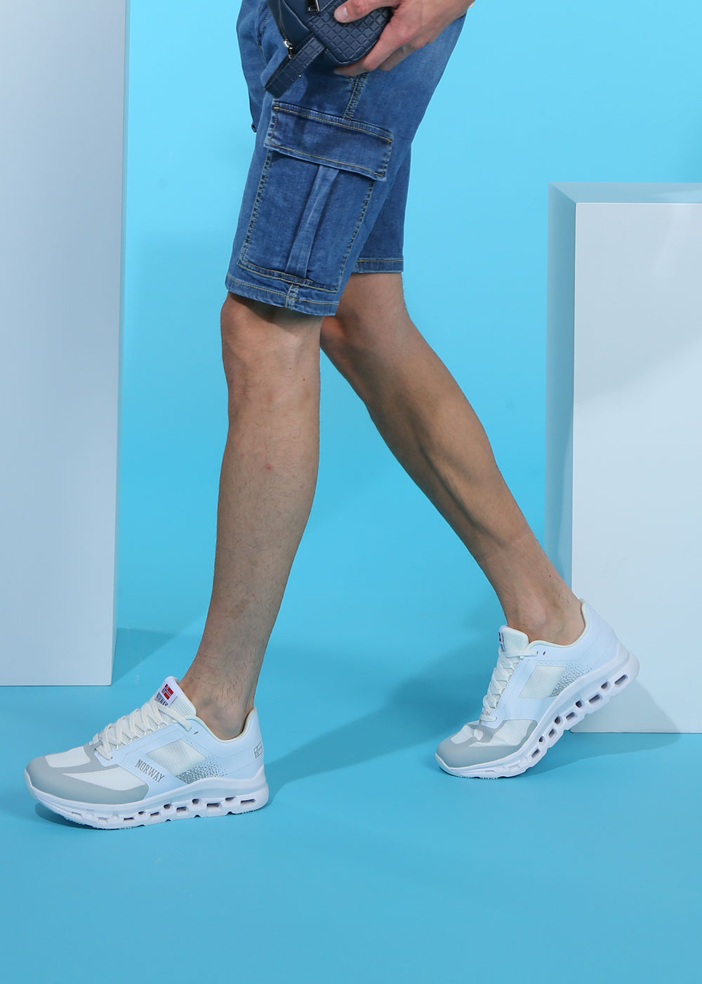 Male model's legs in denim cargo shorts wearing white trainers walking on a blue studio background