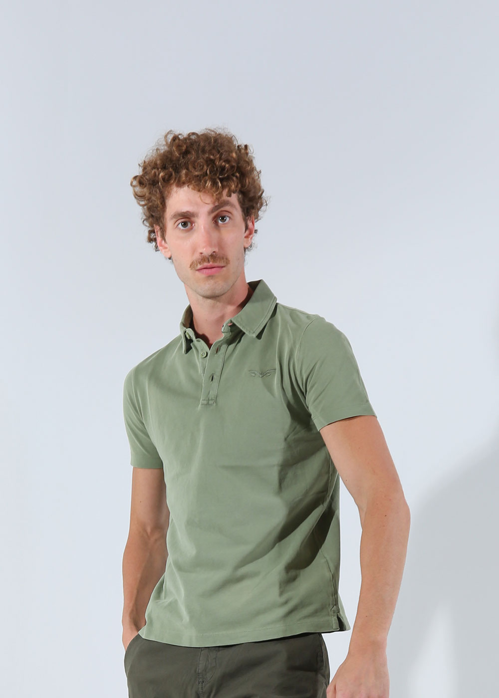 Male model with curly hair and moustache wearing a green polo shirt and dark trousers, standing against a plain light background