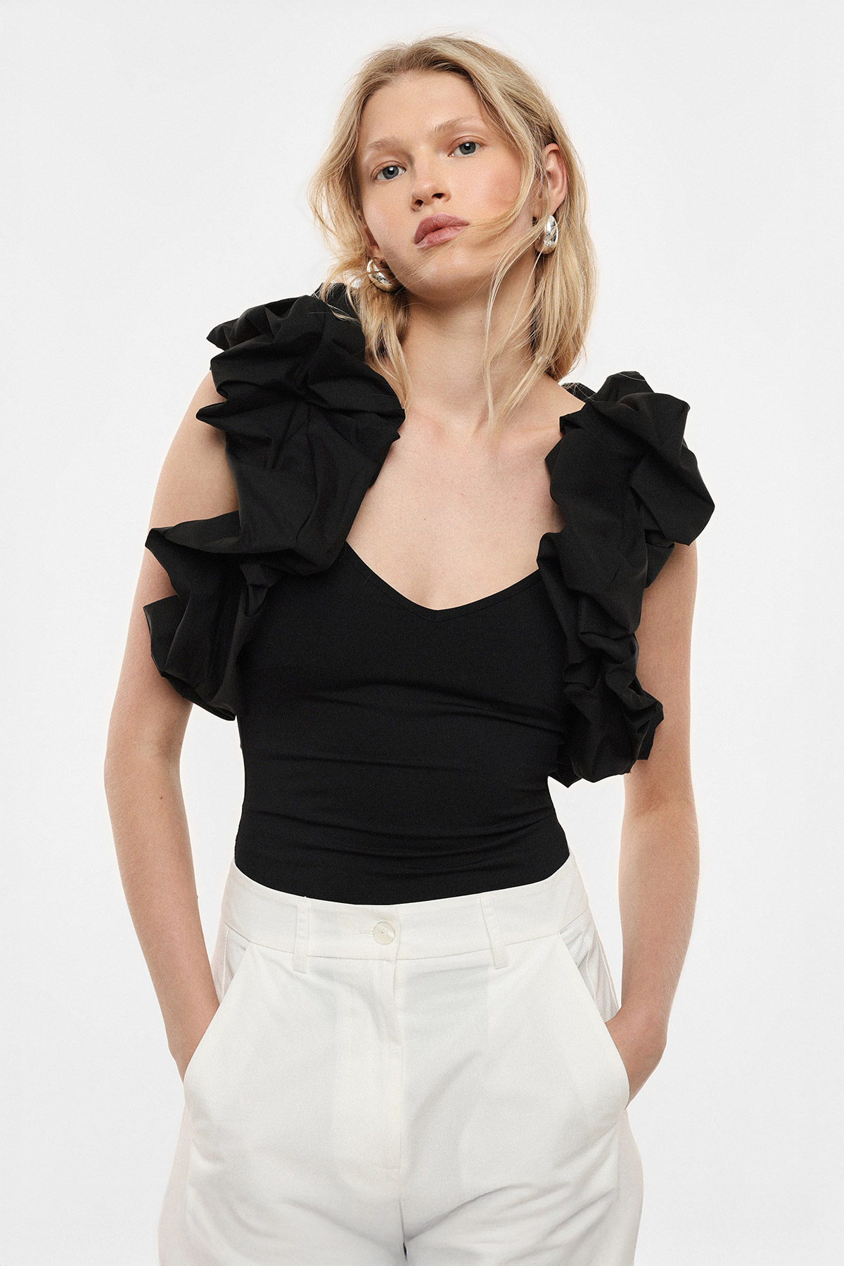 Woman wearing black ruffle-shoulder top and white trousers, hands in pockets, fashion portrait on plain background