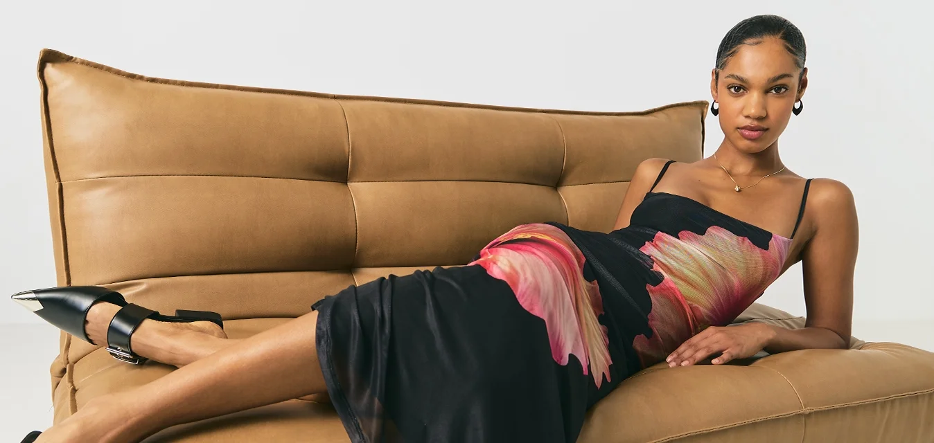 Donna sdraiata su un divano in pelle color sabbia, che indossa un vestito nero con stampa floreale rosa e scarpe nere con punta e cinturini alla caviglia.