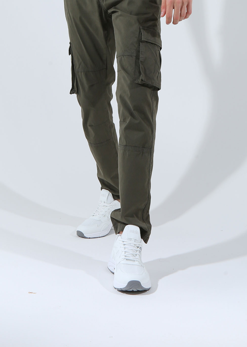 Lower body of model wearing olive-green cargo trousers with side pockets and white trainers on a plain studio background