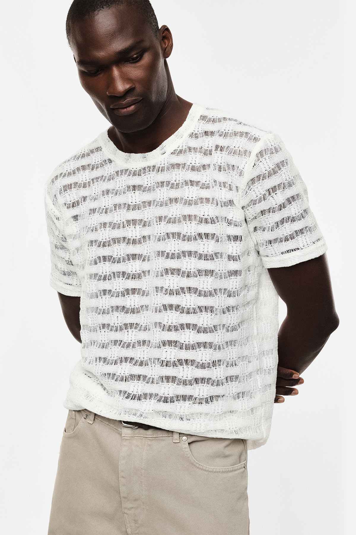 Male model wearing white open-knit striped short-sleeve sweater and beige trousers, hands behind back, looking down