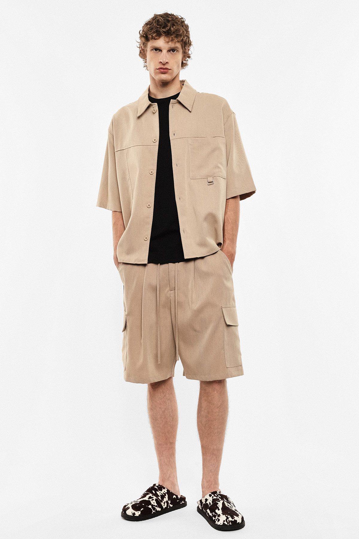 Male model in beige short-sleeve overshirt and cargo shorts with black T-shirt and patterned slip-on shoes on white background