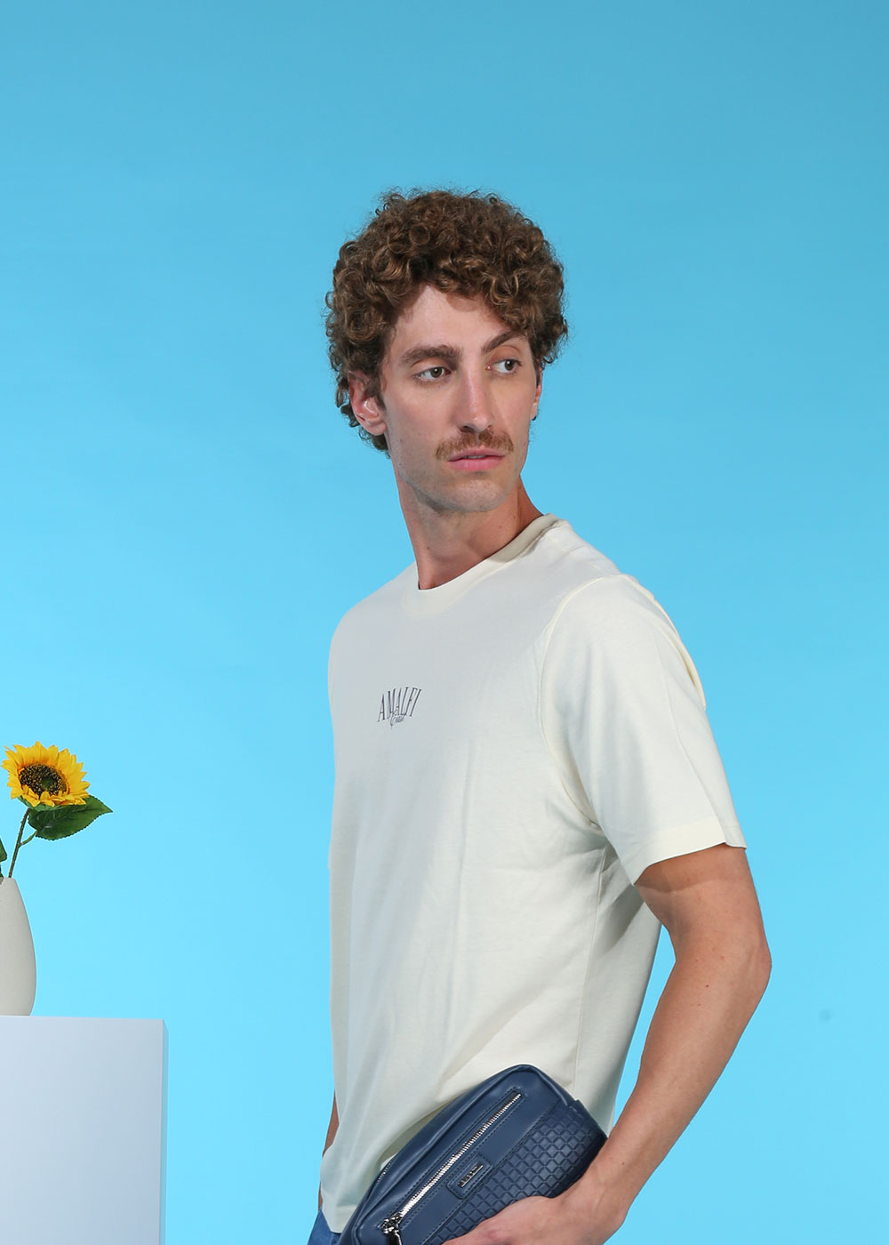 Man with curly hair and moustache wearing a cream T-shirt and holding a navy toiletry bag against a blue background with a sunflower
