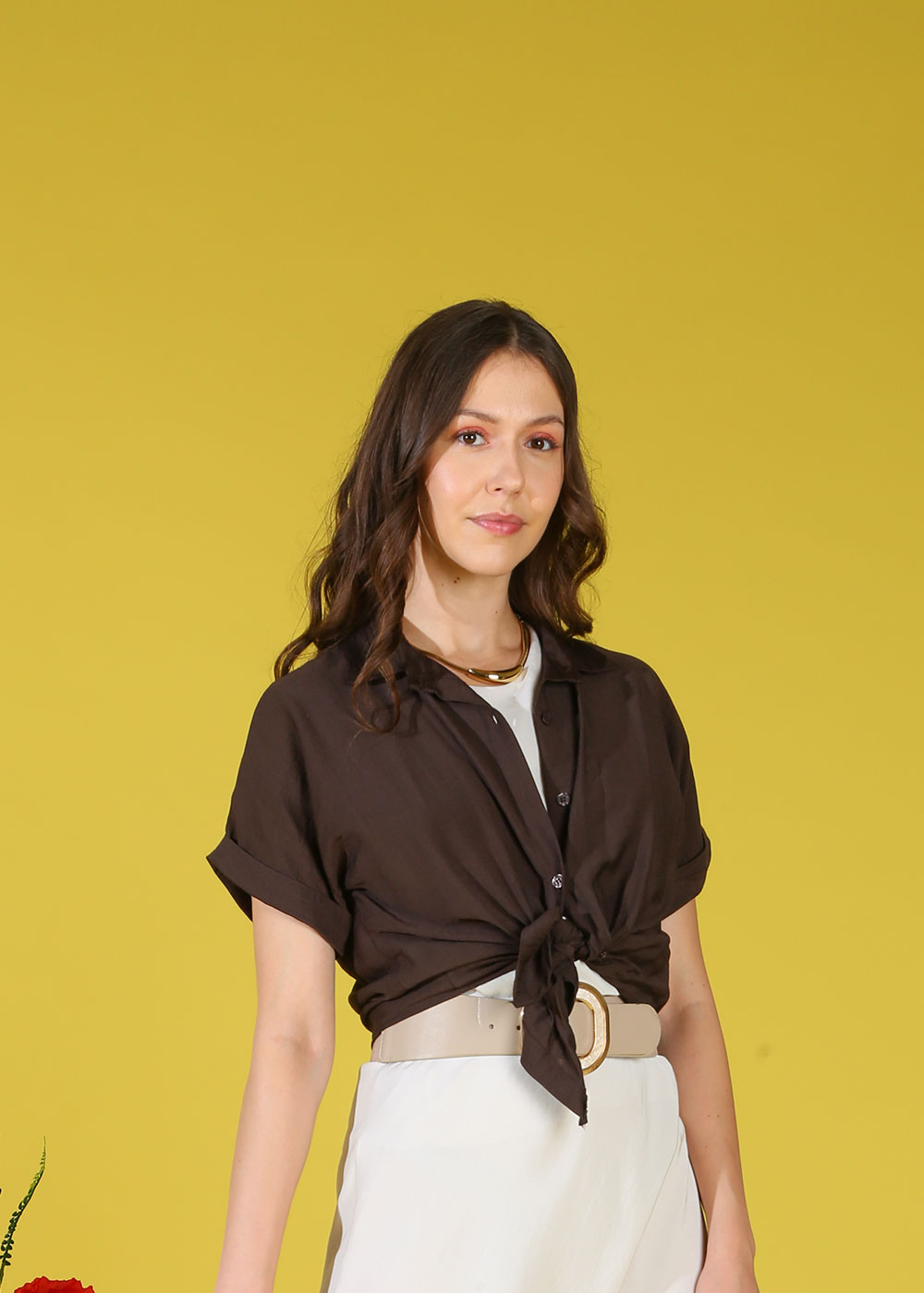 Woman wearing a knotted brown shirt, white skirt and beige belt with a gold necklace, standing against a solid yellow background