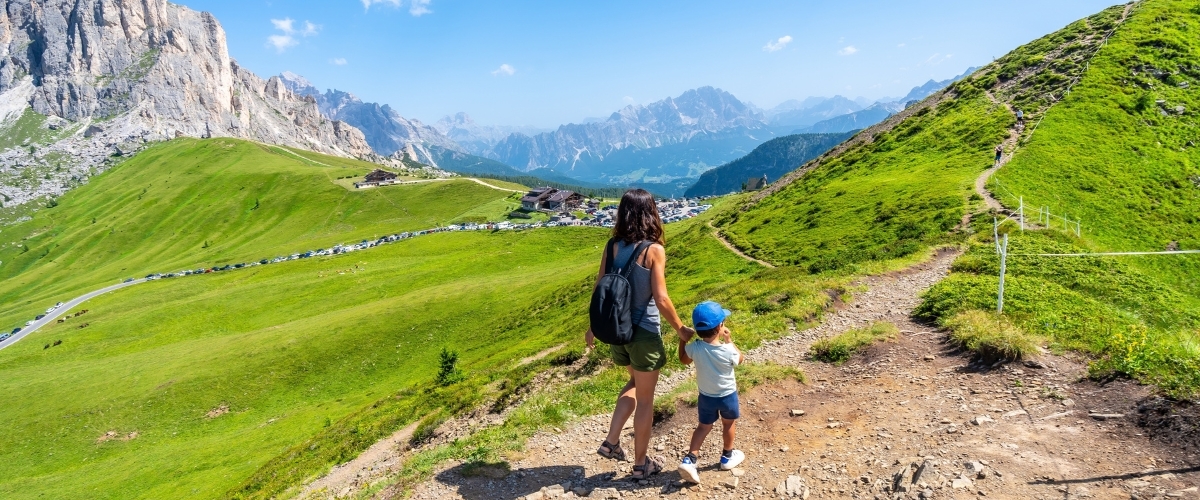 Mother and child hiking on a grassy mountain trail in the Alps, panoramic view of green meadows and rocky peaks