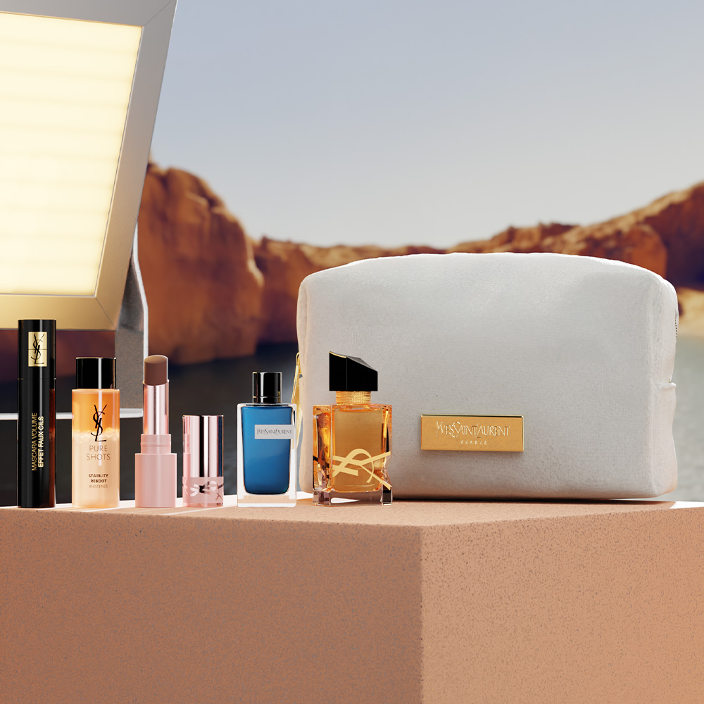 Una selezione di prodotti di bellezza YSL, tra cui profumi, un rossetto, un siero e un mascara, disposti su un blocco di pietra accanto a un beauty case beige.