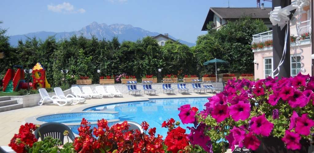 Outdoor hotel swimming pool with sun loungers, bright red and magenta flowers, children's play area and mountains in the background