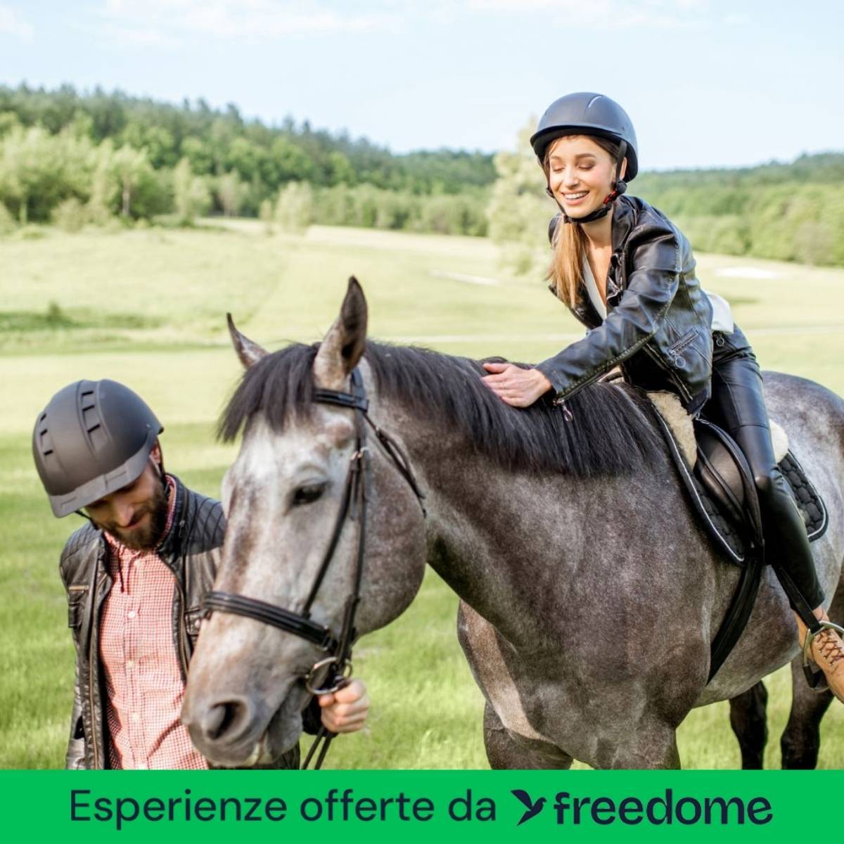 Smiling woman in a helmet riding a grey horse while a man leads it across a sunlit green field, with a 'freedome' banner
