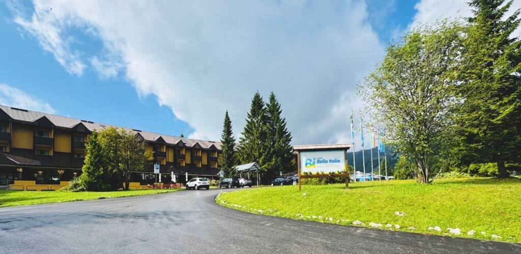 Alpine resort hotel with cars in the forecourt, green lawns and a 'Bella Italia' sign under a cloudy blue sky