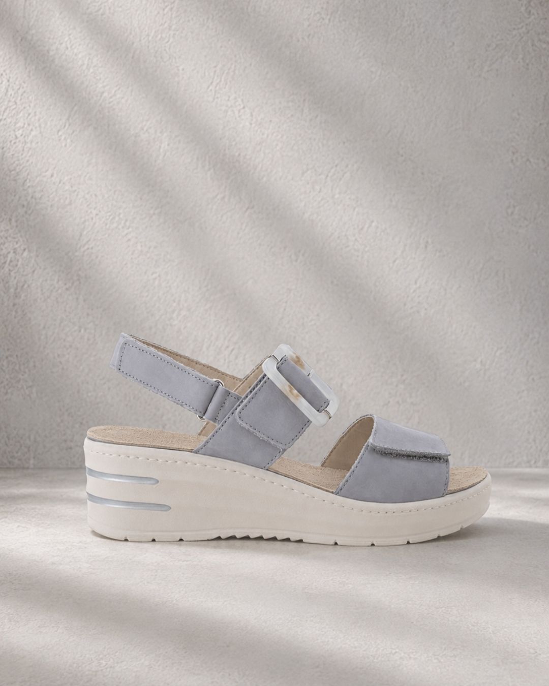 Light gray women's suede wedge sandal with ankle strap and decorative buckle on neutral textured background