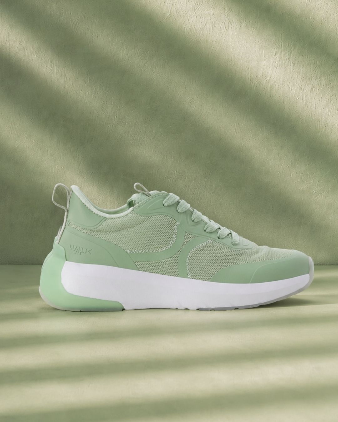Mint green athletic sneaker side view with mesh upper and chunky white sole on matching green backdrop with soft striped shadows