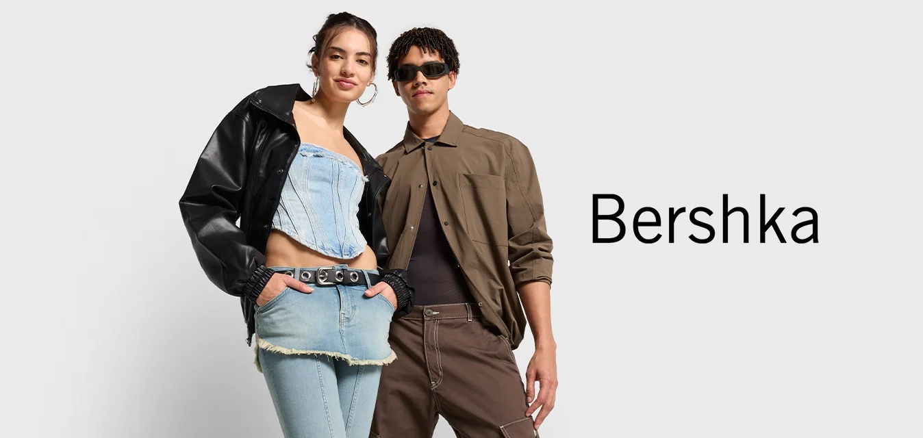 Due persone stanno fianco a fianco. Uno indossa una giacca di pelle nera e un top in denim corta con jeans a vita alta. L'altro indossa una camicia marrone e pantaloni.