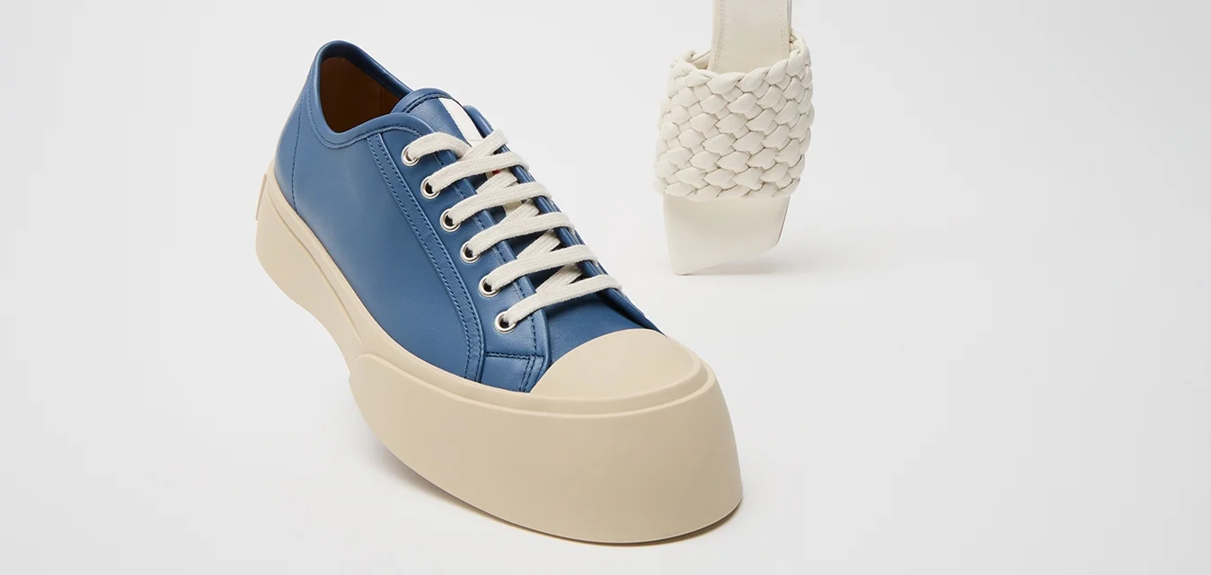 Sneaker in pelle blu con lacci bianchi e una suola spessa beige; accanto, un sandalo a slittamento bianco intrecciato con un tacco curvo.