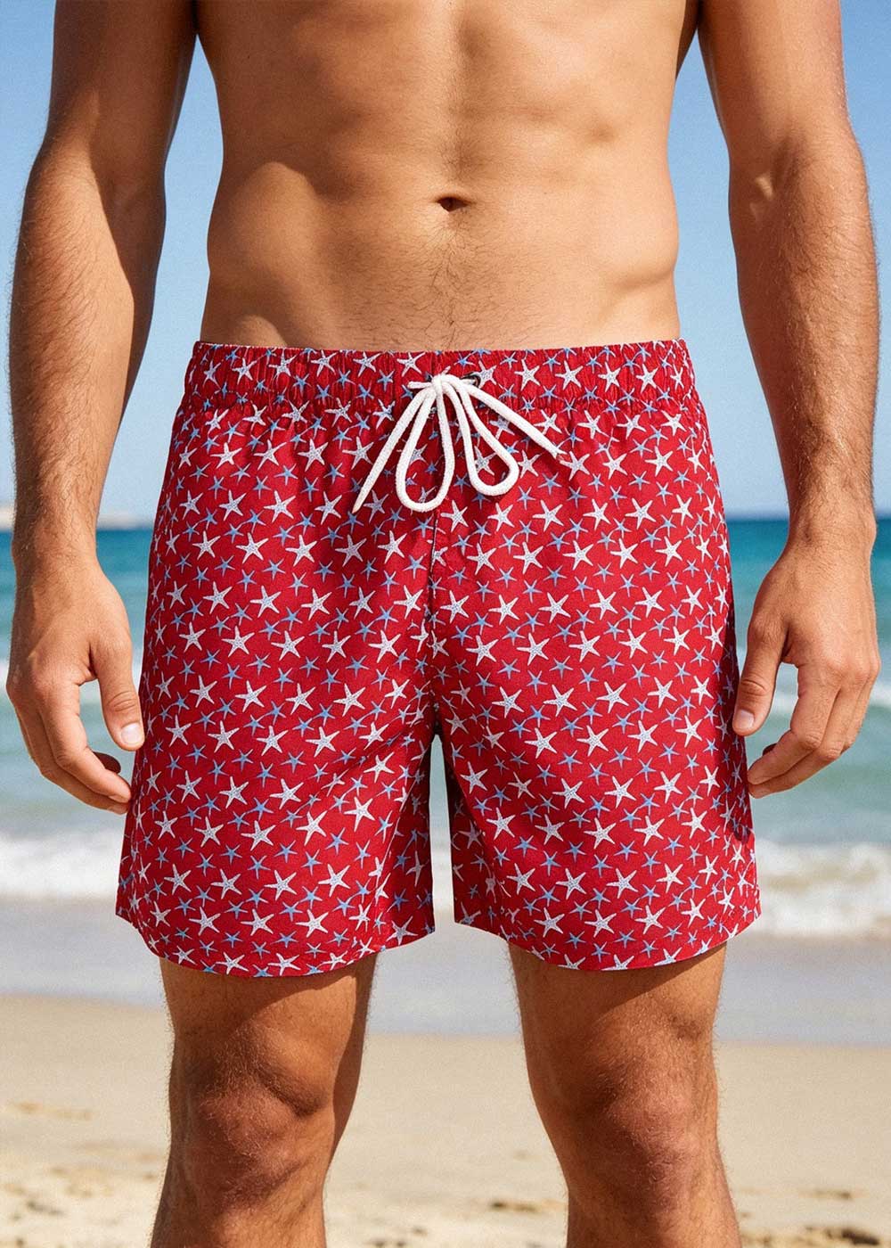 Man's torso and legs wearing red swim shorts with white starfish print and drawstring, standing on a sandy beach by the sea
