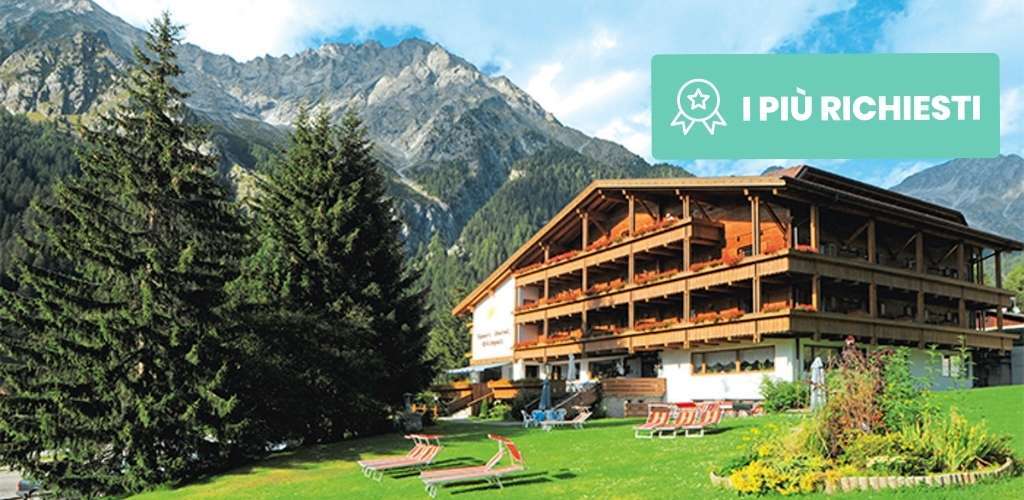 Alpine wooden chalet hotel with balconies, sunbeds on a green lawn, pine trees and rugged mountain peaks under a blue sky