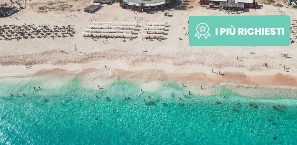 Aerial view of crowded sandy beach with rows of sunbeds, turquoise sea and swimmers; turquoise "I PIÙ RICHIESTI" badge top-right