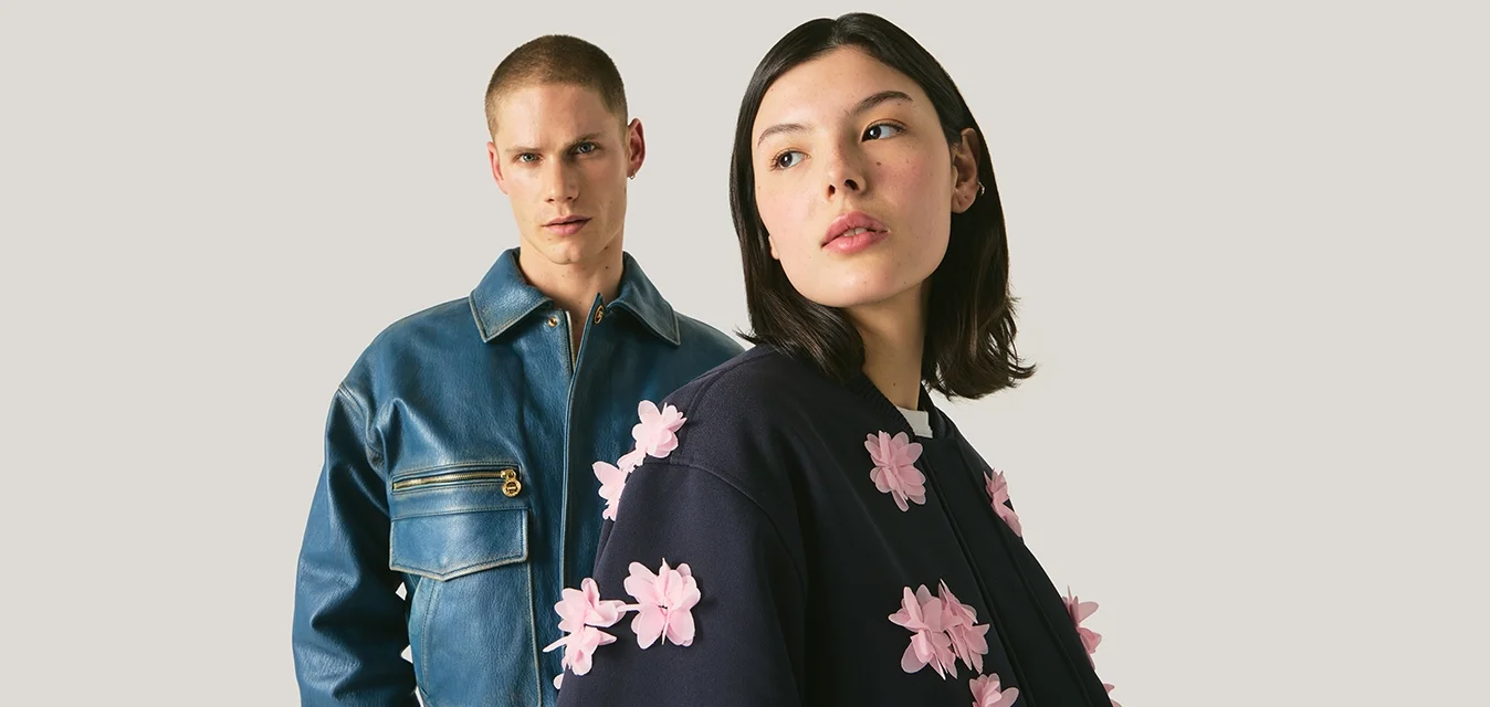 Due modelle indossano giacche alla moda, una in giacca di pelle blu, l'altra in giacca blu scuro con applicazioni floreali rosa in rilievo.