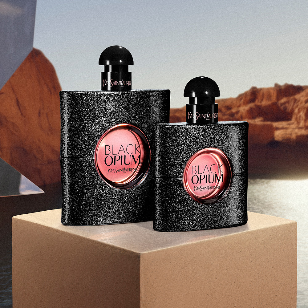 Due flaconi neri brillantinati del profumo YSL Black Opium esposti su un blocco di pietra con un canyon desertico sullo sfondo.