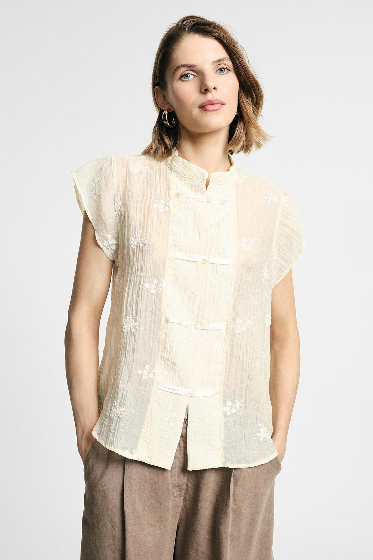 Woman modelling a cream embroidered short-sleeve Mandarin-collar blouse with frog closures and brown trousers