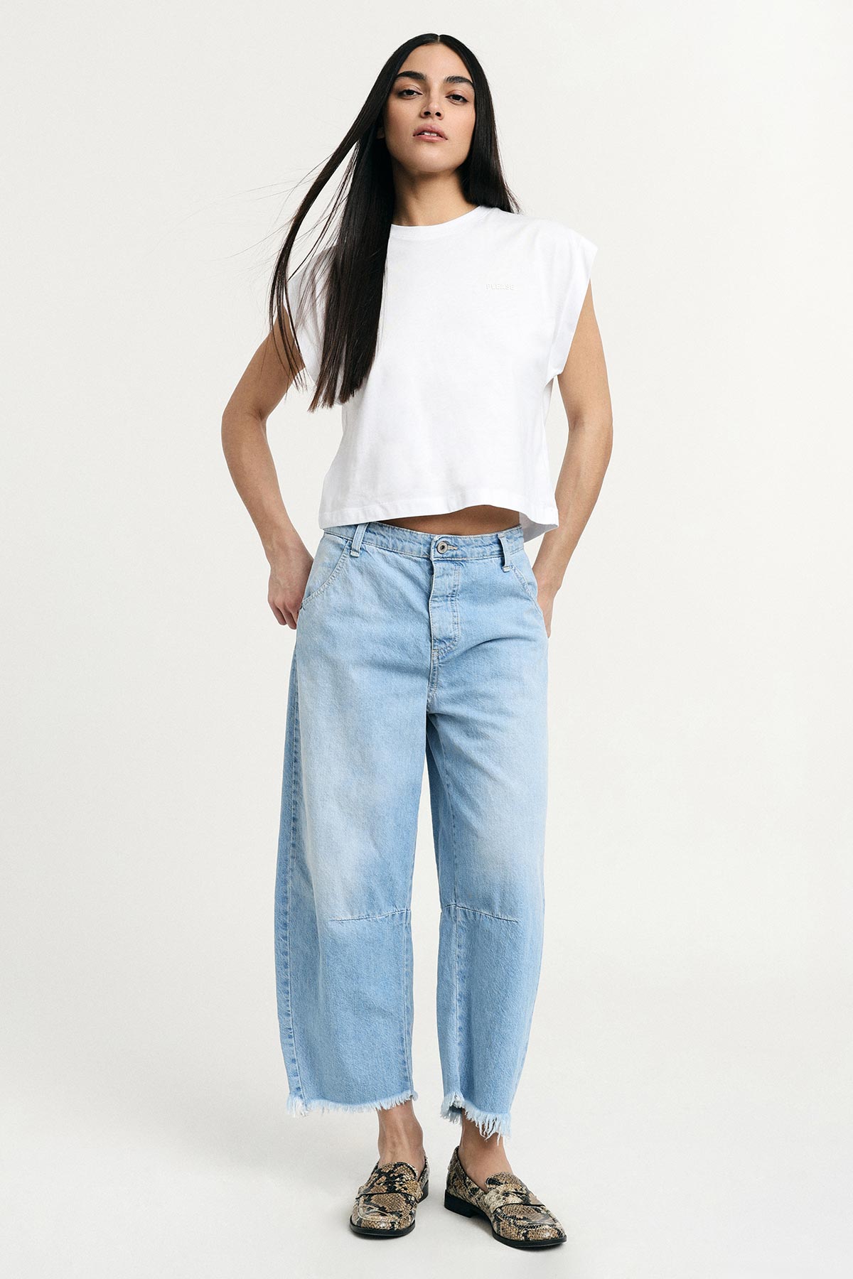 Woman with long dark hair wearing a white sleeveless crop top, light-blue wide-leg cropped jeans with frayed hem and snakeskin loafers