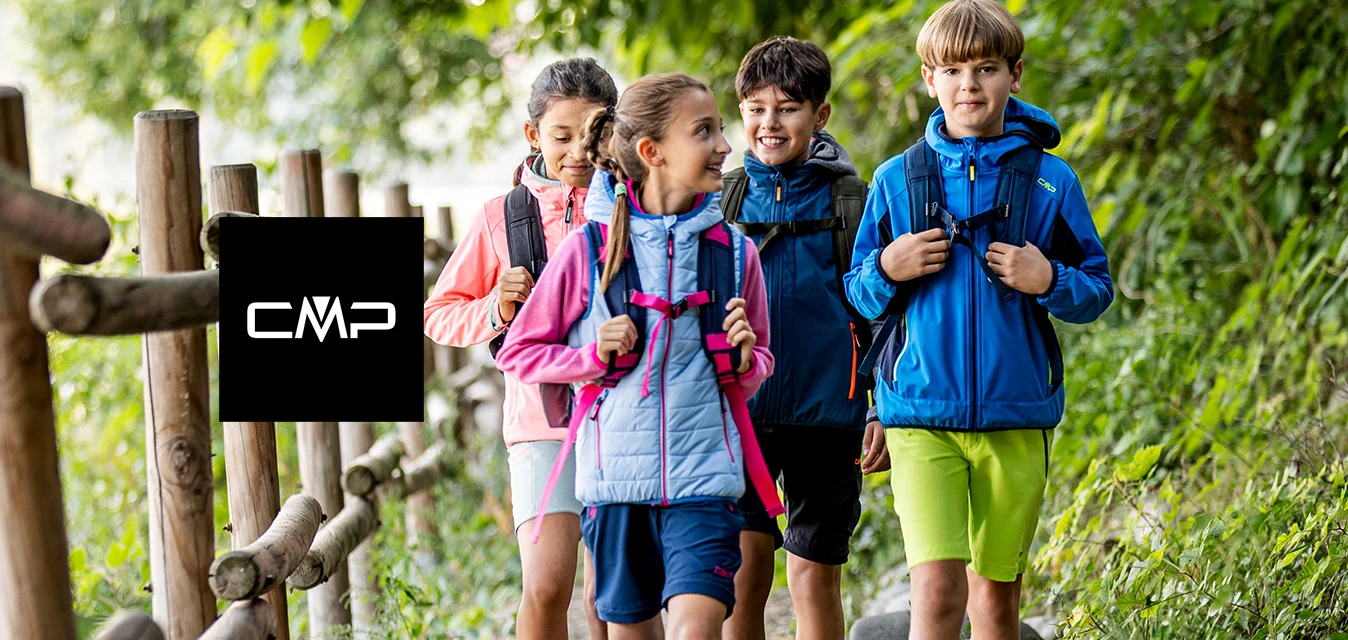 Quattro bambini con zaini che camminano su un sentiero nel bosco accanto a una recinzione di legno, circondati da alberi verdi e fogliame.