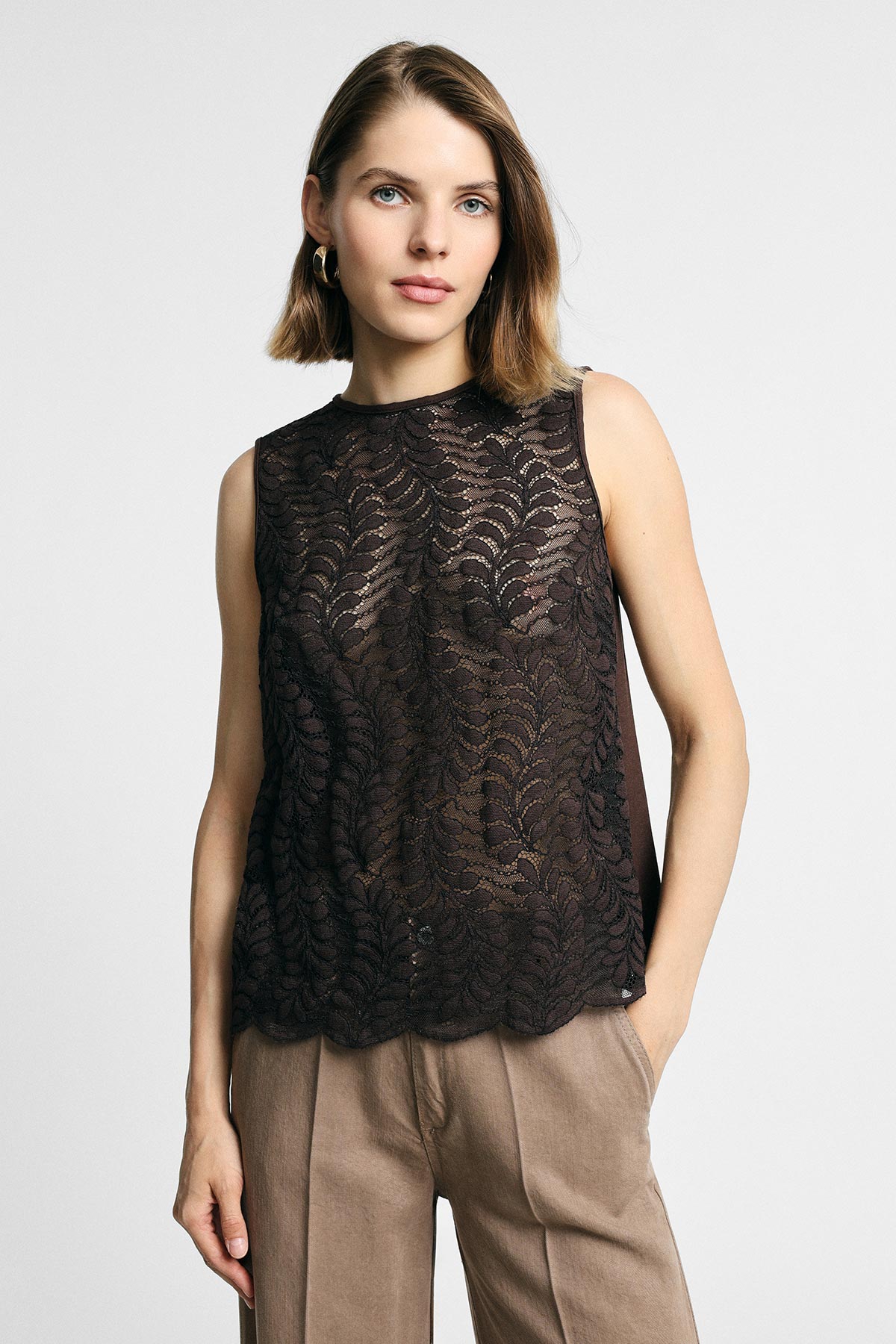 Model wearing a sleeveless black lace blouse and tan high-waisted trousers, standing against a plain light background