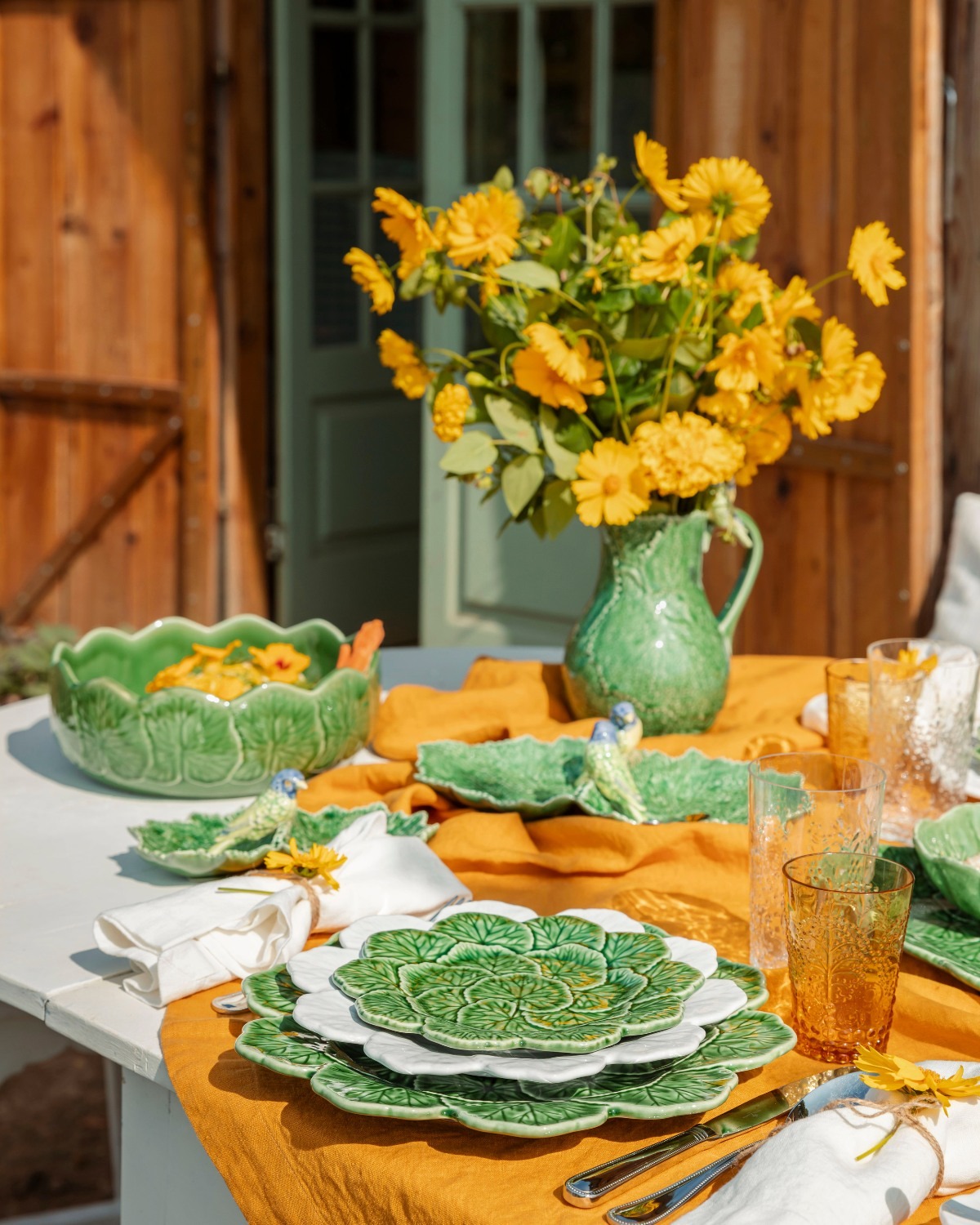 Sunlit outdoor table with green leaf-pattern plates and bowls, mustard cloth, yellow flowers in a green jug and amber glasses
