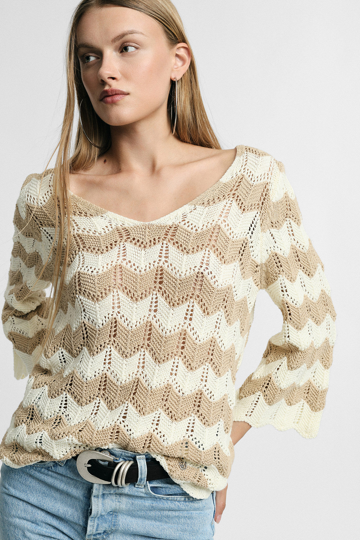 Young woman wearing cream and beige chevron knit V-neck sweater with hoop earrings and light-wash jeans, looking left against plain background