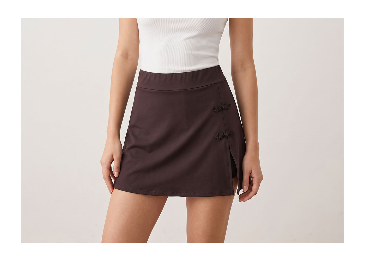 Brown mini skirt with side slit and two decorative frog-knot closures, paired with white ribbed tank, cropped fashion shot