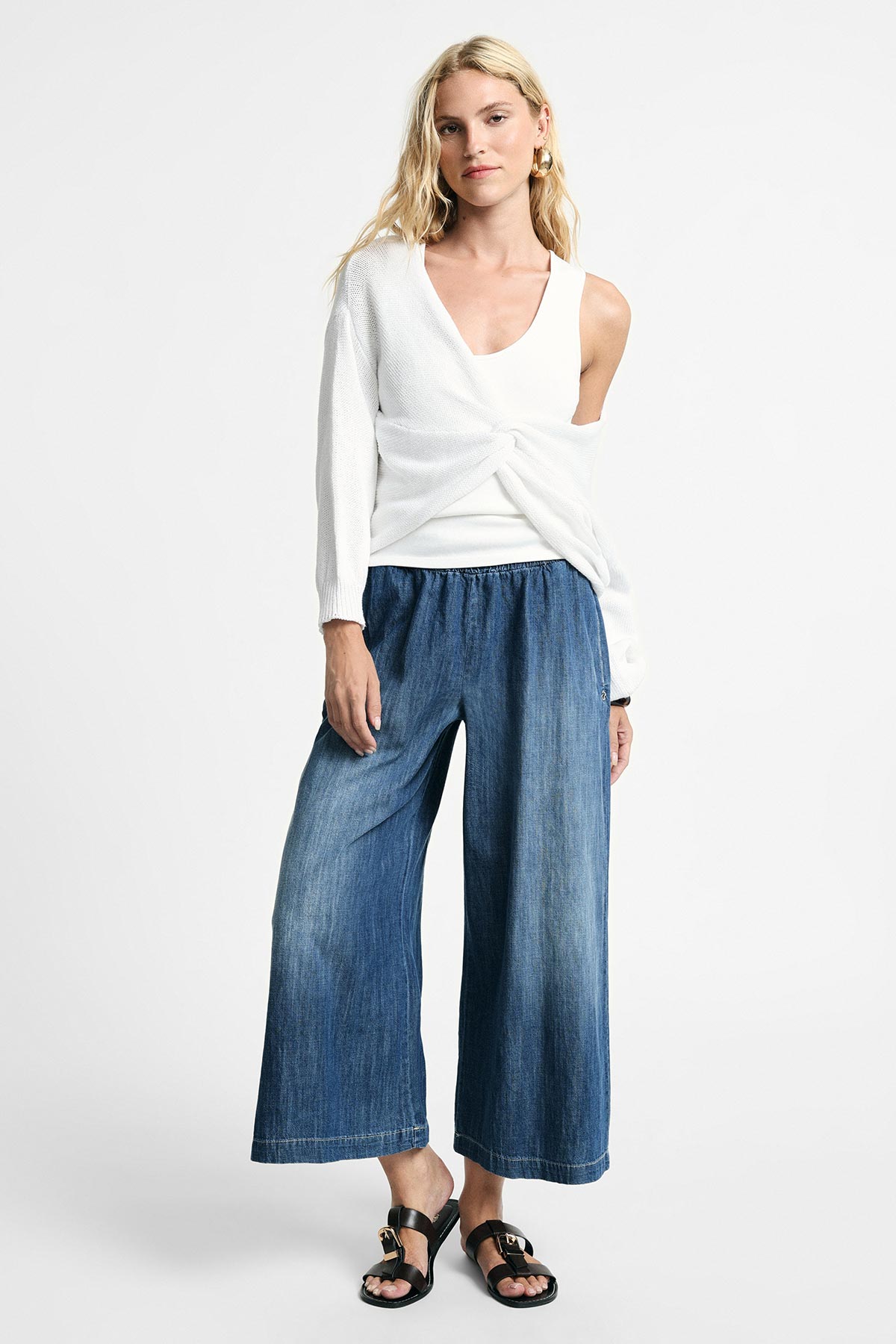 Woman modelling white twisted sweater over white tank top with wide-leg cropped denim trousers and black sandals, studio