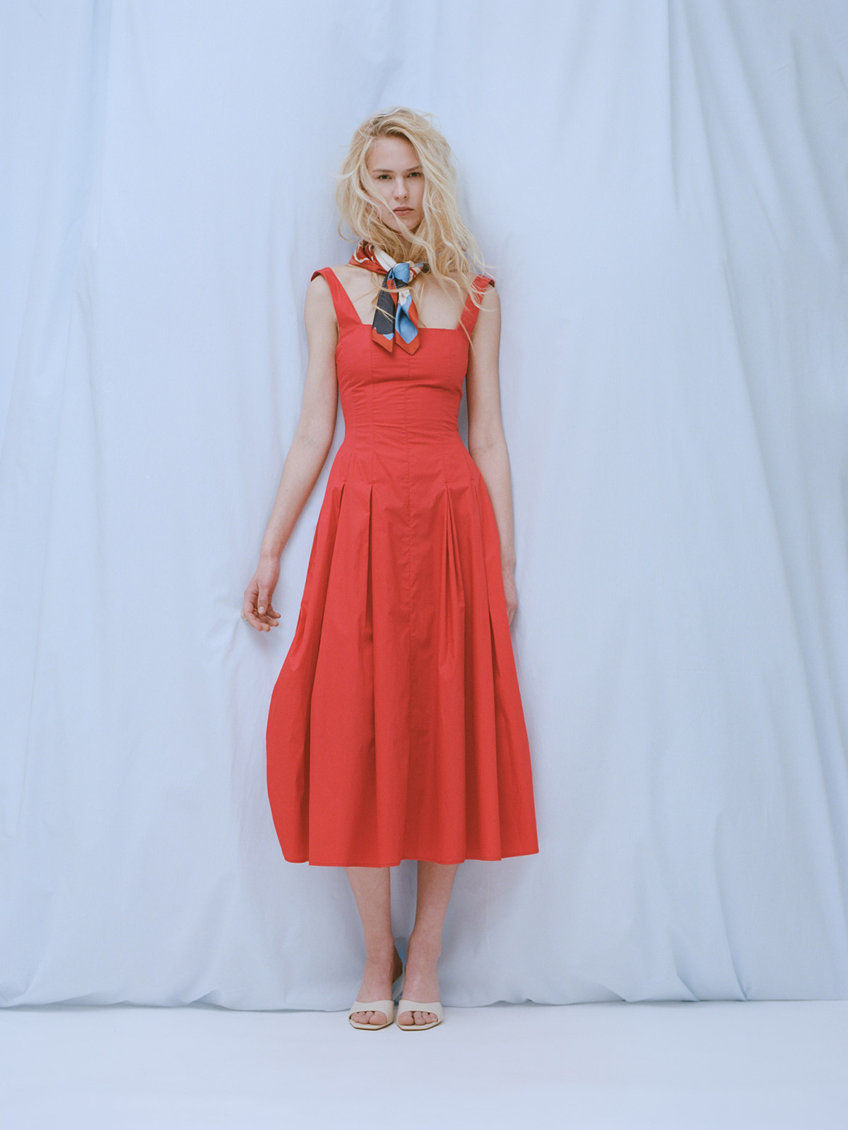 Blonde woman wearing a fitted red midi dress and patterned neck scarf standing against a pale draped backdrop