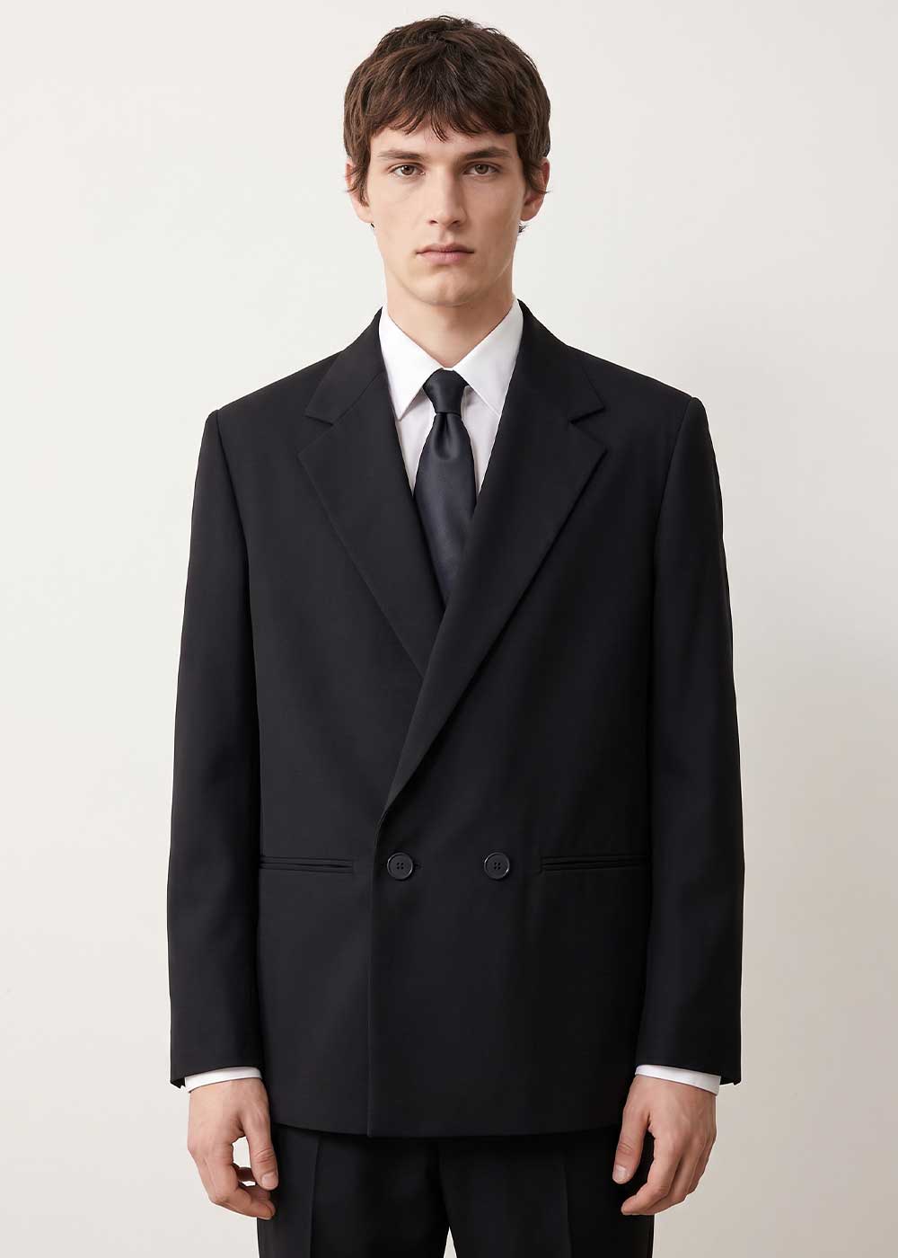 Man in black double-breasted suit with white shirt and black tie standing against a plain white background