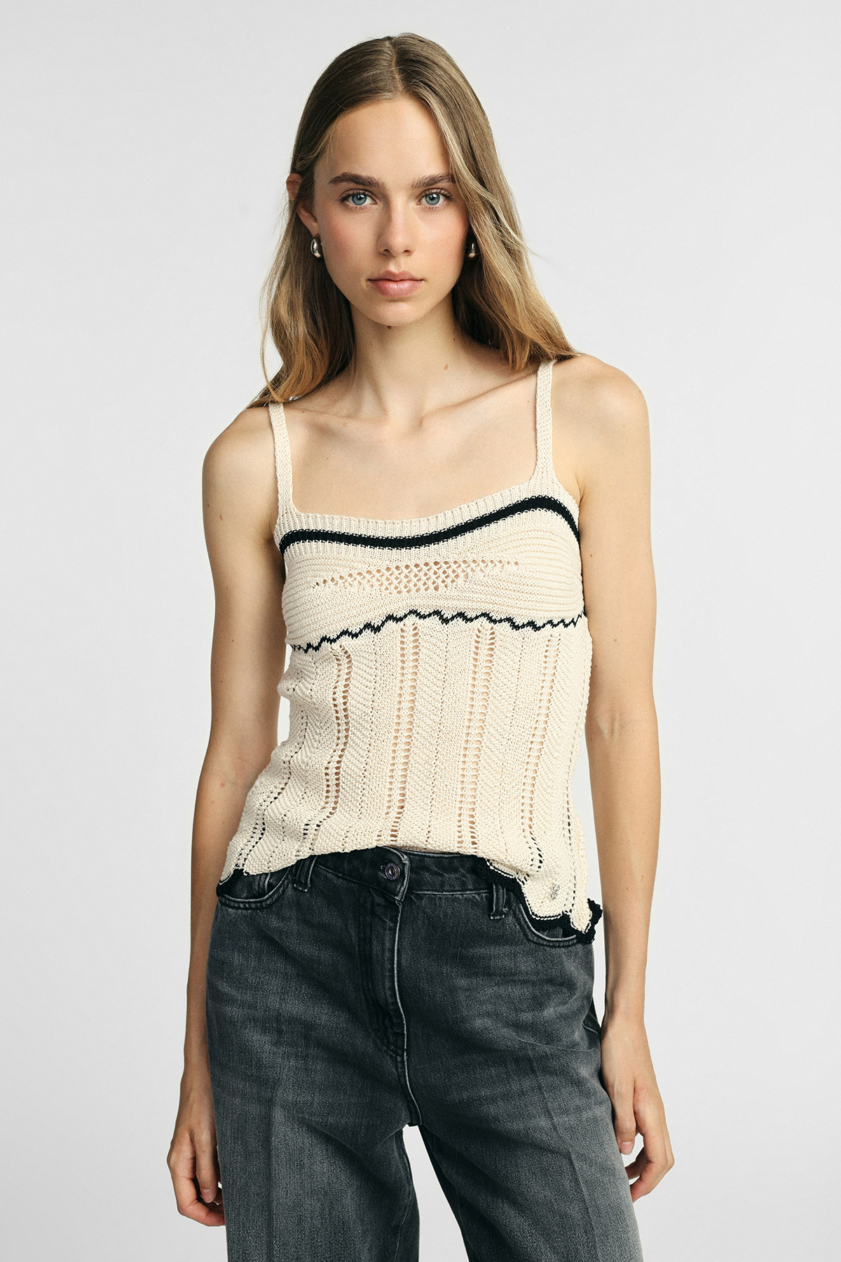 Female model wearing a cream knit camisole with black trim and high‑waisted faded black jeans, standing against a plain light background