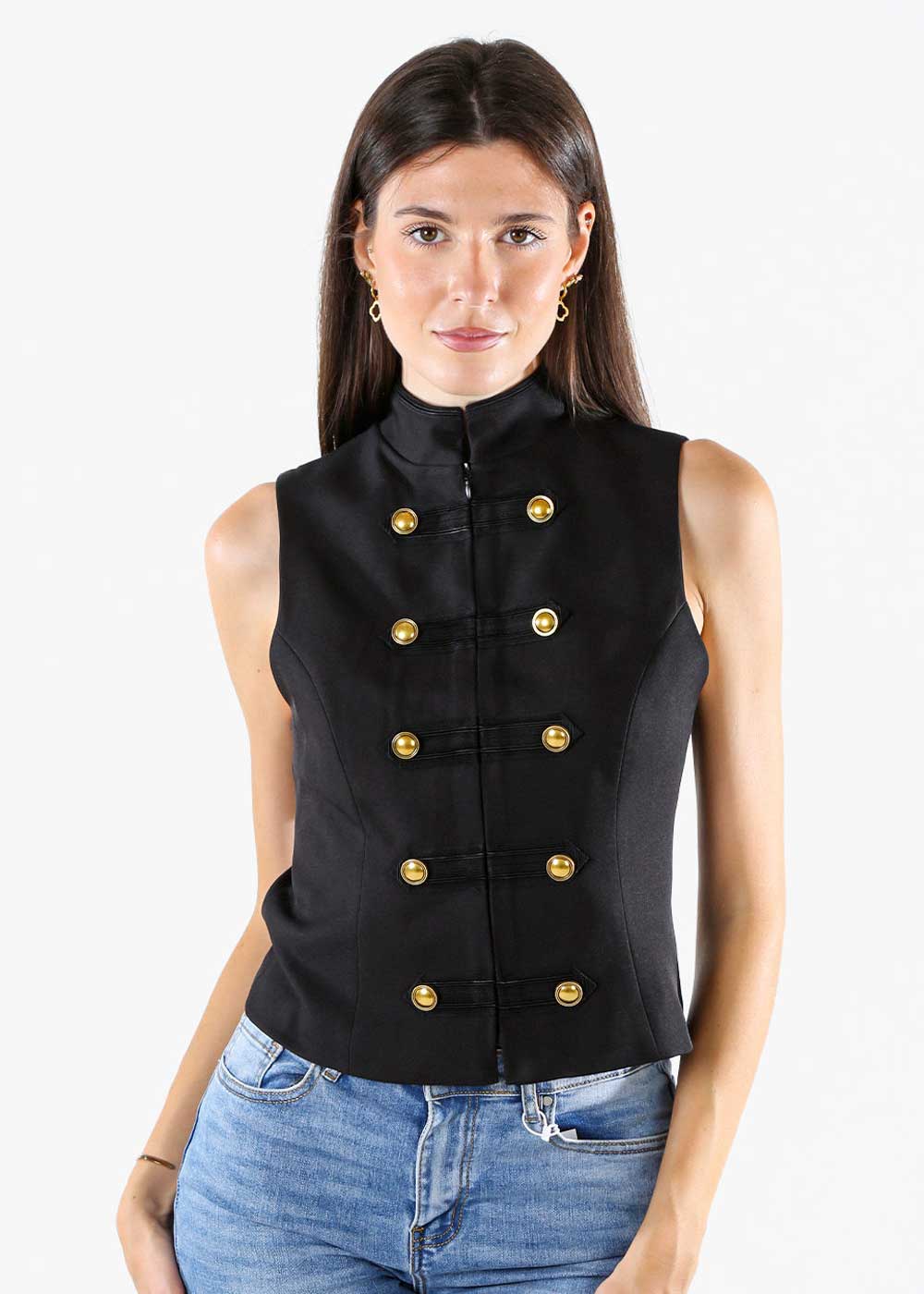 Woman wearing a black sleeveless military-style vest with gold buttons and high collar, paired with blue jeans