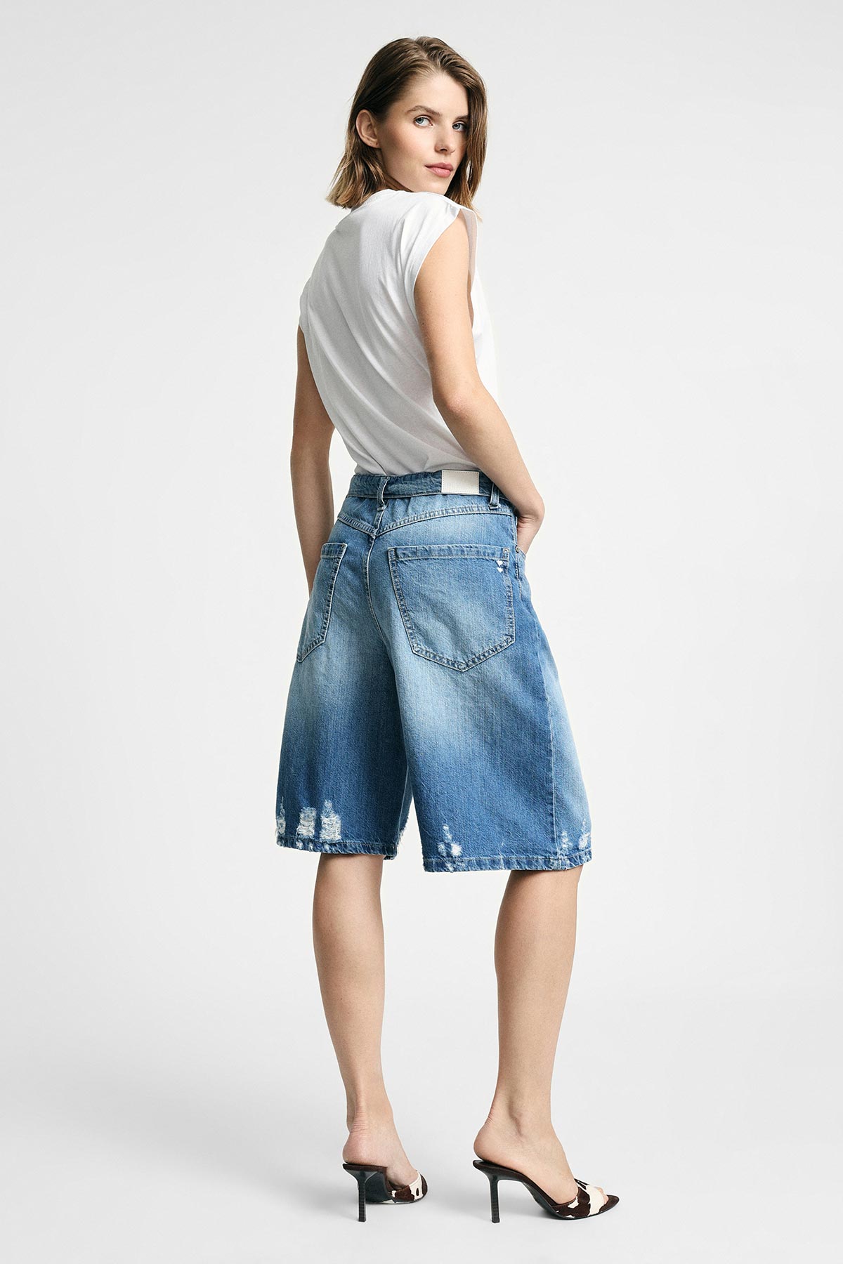 Woman modelling white sleeveless top, distressed knee-length denim shorts and black heeled mules, looking over shoulder