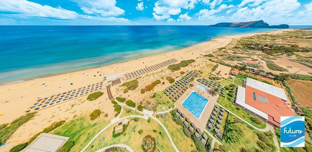 Aerial view of a Mediterranean resort with turquoise sea, long sandy beach lined with sunbeds, outdoor pool, lawns and distant headland