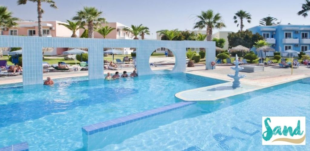 Outdoor resort swimming pool with tiled archway bridge, sunloungers and palm trees by pastel hotel buildings with guests relaxing